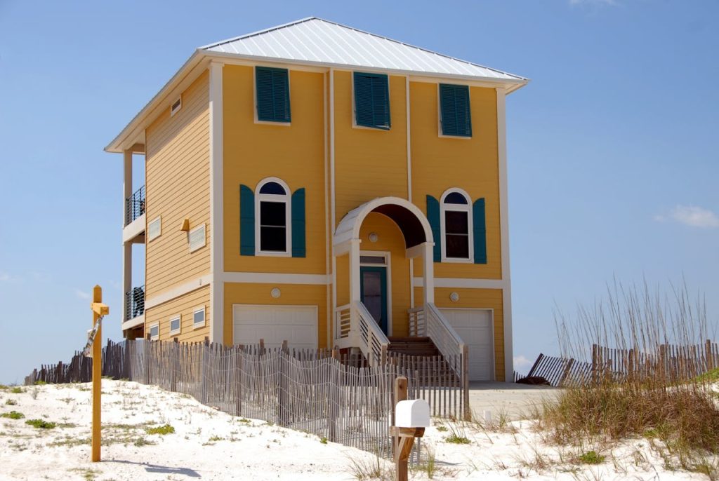 A vibrant yellow beach house with green shutters sits on a sandy dune, surrounded by a fence.